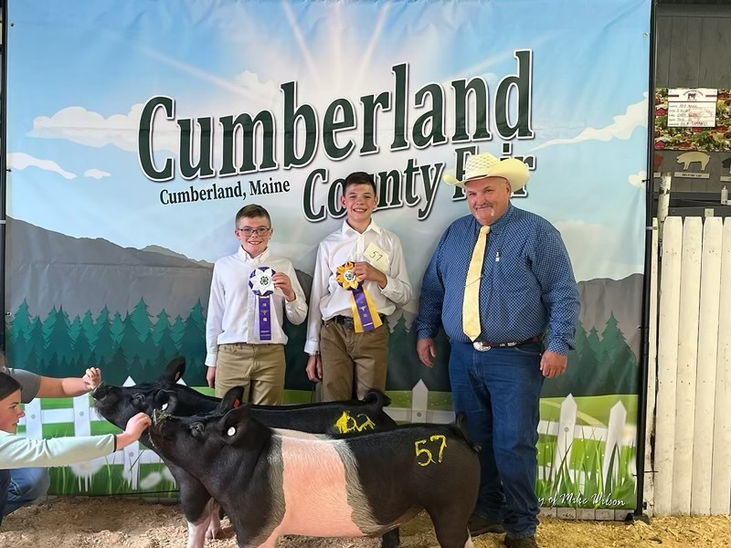 Two students stand in front of a banner for Cumberland County Fair with a judge and their awards and pigs
