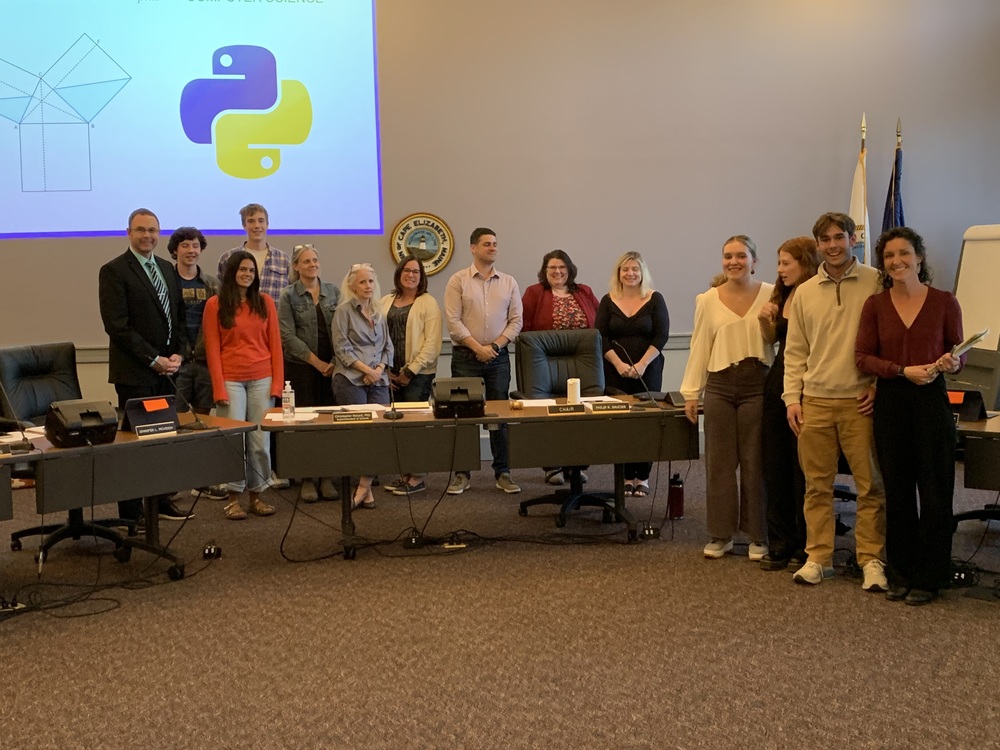 Mrs. Plummer and students stand in front of the school board and Dr. Record, while a slide is reflecting on the wall behind them