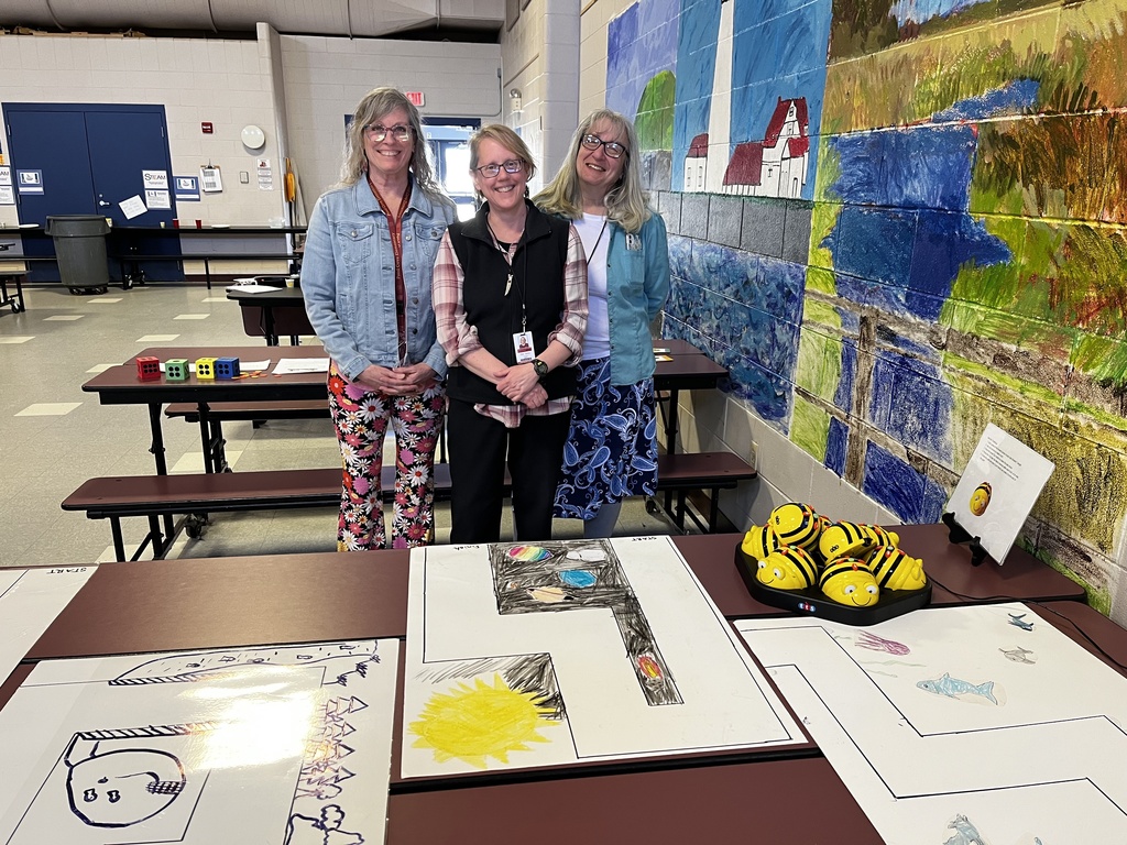 Tami, Ellen, and Leesa stand behind a table with colorful drawings on it next to a very colorful mural that includes a lighthouse