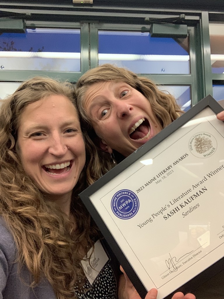 Two women stand with their heads together smiling as one holds an award